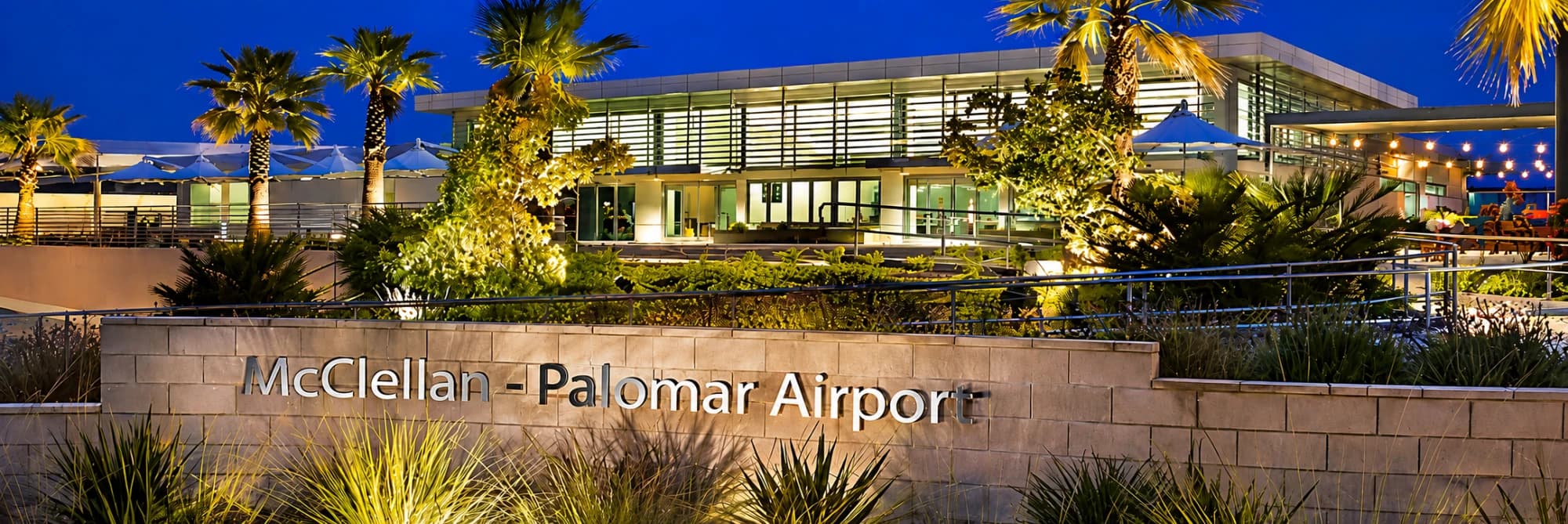 The McClellan-Palomar Airport terminal at dusk, its sign on the stone wall in front, palms lit behind.