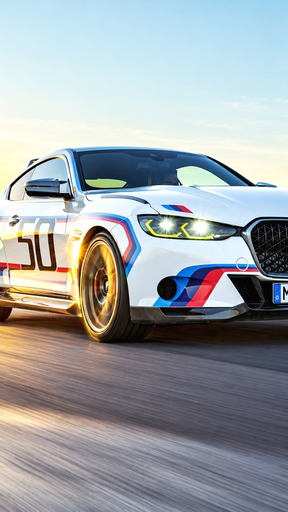 A white BMW coupe in racing livery at speed on a track at sunset.